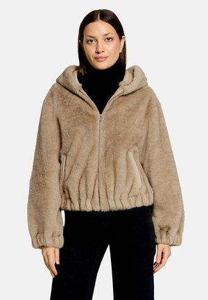 Femme portant une veste à capuche pelucheuse marron clair avec des poignets et un ourlet élastiques, un col roulé noir et un pantalon noir, debout devant un fond uni.