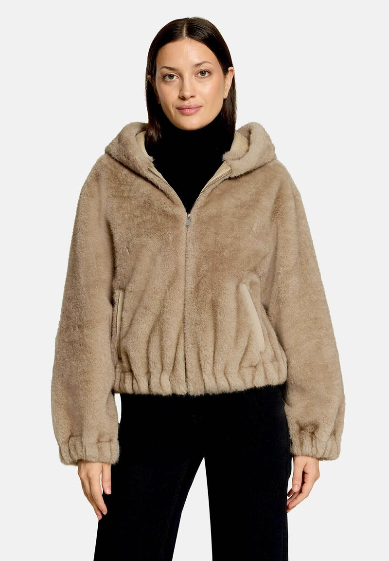 Femme portant une veste à capuche pelucheuse marron clair avec des poignets et un ourlet élastiques, un col roulé noir et un pantalon noir, debout devant un fond uni.