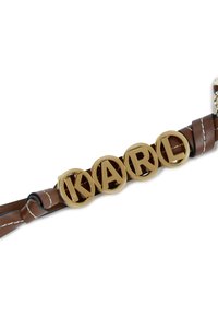 Pruun nahkremm, millel on neli ringikujulist kuldset tähte, mis moodustavad sõna "KARL". Rinnet aitavad kaunid valged õmblused ja hõbedane kettühendus.