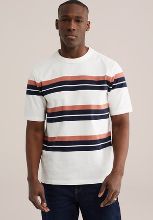 Mees, kes kannab valget lühikese varrukaga särki, millel on horisontaalsed laigud tumesinise ja roostepunase toonis, koos tumedate siniste teksapükstega, seisab ühevärvilise tausta ees.