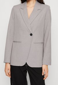 Harmaa bleiseri, joka on valmistettu sileästä kankaasta ja jossa on notched-lapeli, yksinkertainen nappisulku ja kaksi etutaskua, joissa on hienovaraiset ommellut viivat.