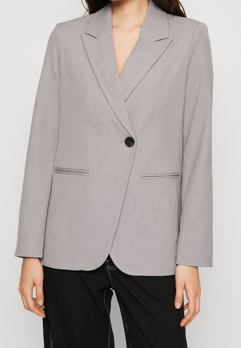 Harmaa bleiseri, joka on valmistettu sileästä kankaasta ja jossa on notched-lapeli, yksinkertainen nappisulku ja kaksi etutaskua, joissa on hienovaraiset ommellut viivat.