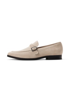 Beżowe mokasyny z zamszu z niskim obcasem, szpiczastym noskiem, plecionym paskiem i srebrną klamrą po boku na białym tle.