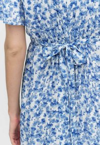 Kukallinen mekko, jossa on sinivalkoinen kuvio, lyhyet hihat, kerätty vyötärö ja solmitys takana. Kevyt kangas.