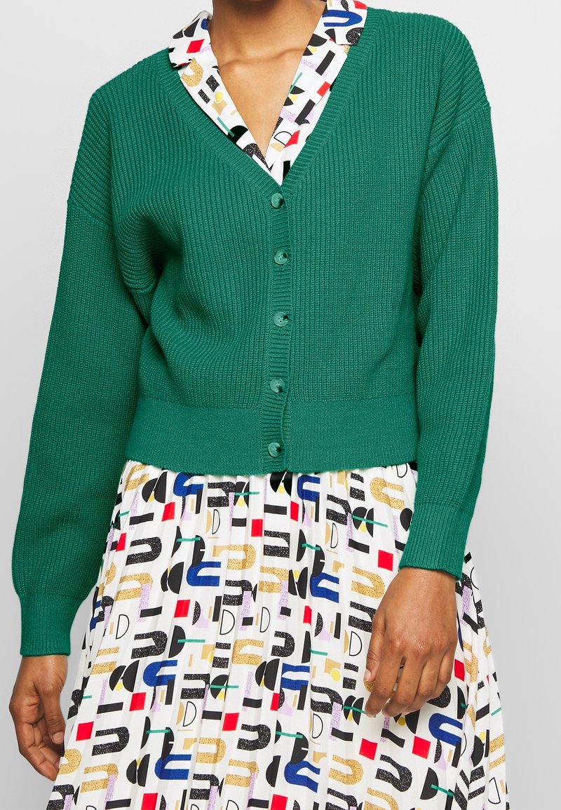 Grön räfflad kofta med knäppning och långärmar, bärs över en flerfärgad mönstrad klänning med abstrakta former och mönster.