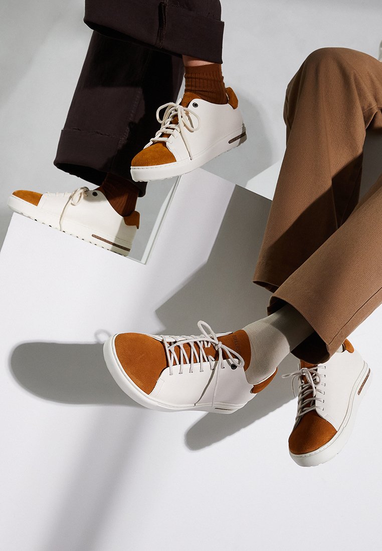 Weiße Sneaker mit Akzenten aus tanfarbigem Wildleder, runde Zehenform, Schnürdesign und einer strukturierten Gummisohle. Auf kontrastierenden Oberflächen präsentiert.