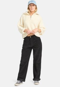 Pull en polaire crème avec col zippé mi-hauteur, associé à un pantalon en velours côtelé noir et des baskets grises. Une casquette bleu clair complète la tenue.
