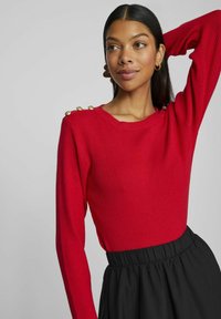 Röd ribbad långärmad topp med guldknappdetaljer på axeln, parat med en svart högmidjad kjol. Slät textur, figurnära design.