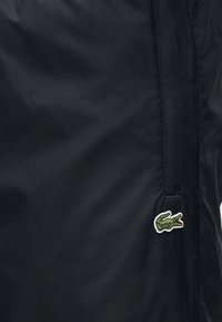 Μαύρο νάιλον μπουφάν με φερμουάρ σε τσέπη και μικρό κεντητό πράσινο λογότυπο κροκοδείλου. Ομαλή υφή με διακριτική γυαλάδα.