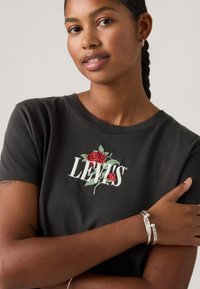 Camiseta negra de algodón con el logo blanco "LEVI'S" y bordado de rosa roja. Presenta un ajuste relajado con mangas cortas. Brazaletes plateados adornan la muñeca.
