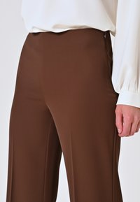 Pantaloni marroni a vita alta con una texture liscia, leggermente elasticizzati e dalla vestibilità sartoriale. La mano mostrata sta tenendo la vita.