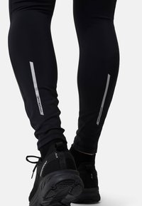 Svarta sportleggings med reflekterande silverränder, tillverkade av stretchigt material. Vristen har en ribbad textur med svarta träningsskor nedanför.