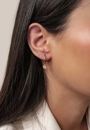Close-up of a woman's ear with straight dark hair wearing a small gold hoop earring featuring a smiling face charm.