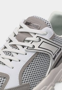 Λευκό και γκρι sneaker Just Cavalli με πλευρές από δίχτυ, λευκά κορδόνια και λογότυπο της μάρκας στη γλώσσα και στην πλευρική επιφάνεια.