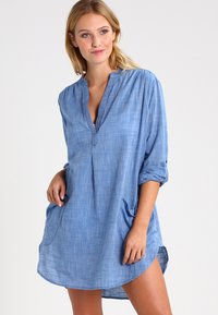 Light blue, woven tunic dress with a V-neck, rolled sleeves, and two side pockets. Soft texture with a casual, relaxed fit.