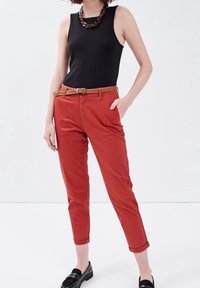 Femme portant un haut noir sans manches, une ceinture tressée marron, un pantalon rouille retroussé aux chevilles et des chaussures noires à enfiler, se tenant avec une main dans la poche.