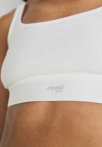 Bijela bešavna braleta s glatkom, rastezljivom teksturom. Ima mekan brandirani elastični pojas s natpisom "sloggi" u svijetlo sivoj boji.