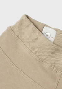 Beige Baumwollstoff mit einem gerippten Bund. Verfügt über ein weißes Etikett mit einem Knopf und sauberen Nähten entlang der Säume.