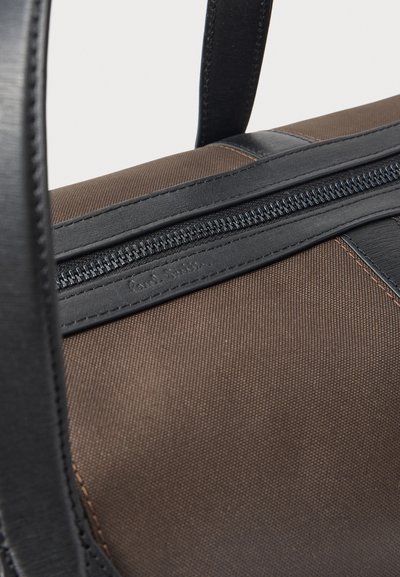 Καφέ υφασμάτινη τσάντα με μαύρες δερμάτινες λαβές και διακόσμηση, με μαύρο φερμουάρ που εκτείνεται οριζόντια στην κορυφή.