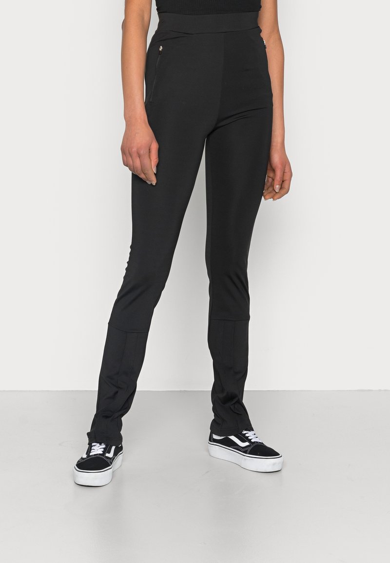 Leggings noirs extensibles avec taille haute, dotés de poches latérales zippées et de jambes fuselées, associés à des baskets plates noires.