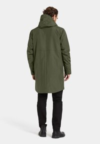 Groene waterdichte jas met capuchon, met een lengte tot halverwege de dijen en een rechte snit. Draagt over een zwarte broek en zwarte laarzen.