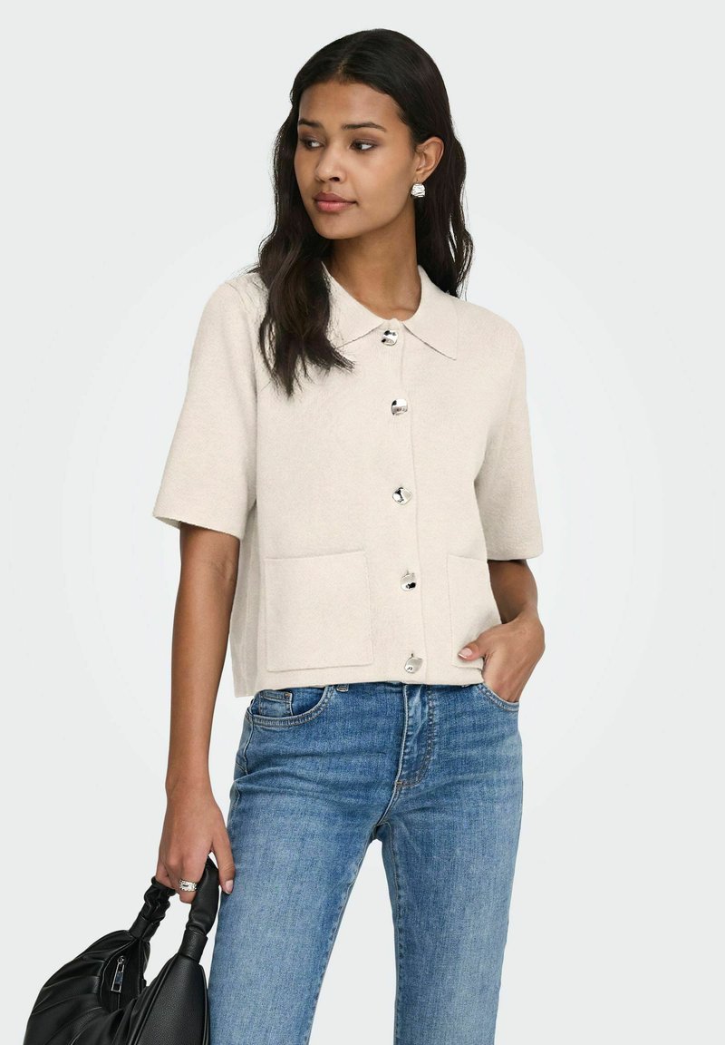 Femme portant un cardigan crème à manches courtes boutonné et un jean bleu, tenant un sac à main noir, une main dans la poche, regardant sur le côté.