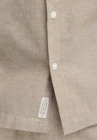 Chemise en lin beige avec des boutons blancs et une petite étiquette d'entretien du tissu cousue près de l'ourlet inférieur.