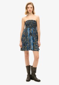 Femme portant une robe bleue à motifs sans bretelles avec une ceinture à pampilles et des bottes en cuir noir mi-mollet, debout devant un fond blanc uni.