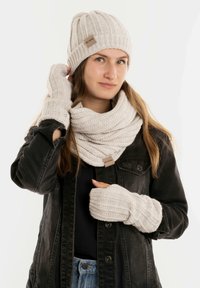Beige gebreide muts, sjaal en vingerloze handschoenen met ribbelstructuur; heeft een klein merklabel op de sjaal; gedragen met een zwarte jas.