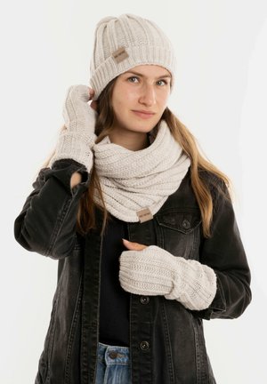 Beige gebreide muts, sjaal en vingerloze handschoenen met ribbelstructuur; heeft een klein merklabel op de sjaal; gedragen met een zwarte jas.