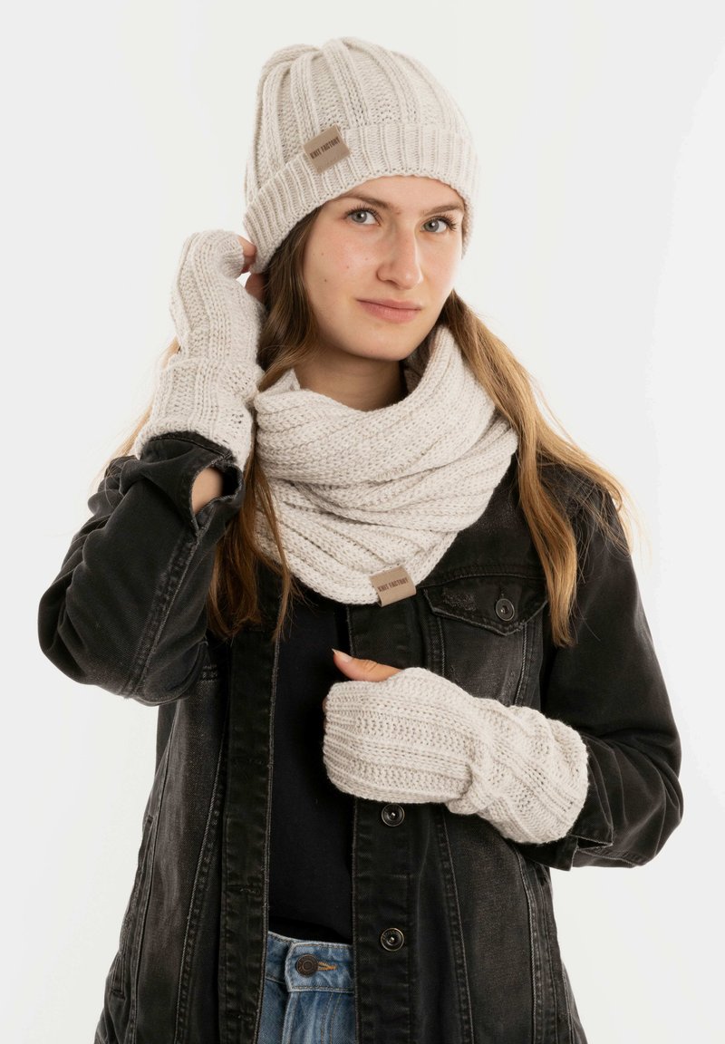 Beige gebreide muts, sjaal en vingerloze handschoenen met ribbelstructuur; heeft een klein merklabel op de sjaal; gedragen met een zwarte jas.