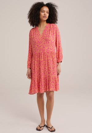 Rochie roz cu model floral, decolteu în V, mâneci lungi și fustă în straturi, realizată dintr-un material ușor. Purtată cu sandale negre fără toc.
