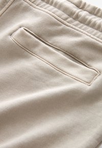 Tessuto in cotone beige con una leggera trama, dotato di una tasca posteriore con cuciture decorative e una cintura elastica.