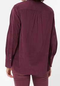 Chemise en velours côtelé bourgogne à manches longues avec un poignet boutonné, détails de pli au dos et un ourlet arrondi. Tissu texturé avec des rayures verticales.