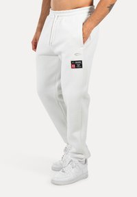 Pantalones de chándal blancos de algodón con cintura ajustable, bolsillo lateral y acento de etiqueta negra. Combinados con zapatillas blancas.