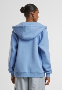 Felpa con cappuccio di colore azzurro chiaro, tessuto morbido, vestibilità rilassata, tasca a marsupio, polsini e bordo a coste, dettagli di cucitura visibili, indossata con pantaloni grigi chiari.