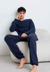 Marineblauwe lange mouwen top en gemêleerde pyjamabroek in donkerblauw met rode accenten, zittend op een lichtgekleurde, gestructureerde stoel.