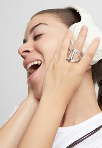 Anillo de plata con diseño puntiagudo en una mano, auriculares blancos de diadema en la cabeza de una mujer, que lleva una camiseta blanca lisa.