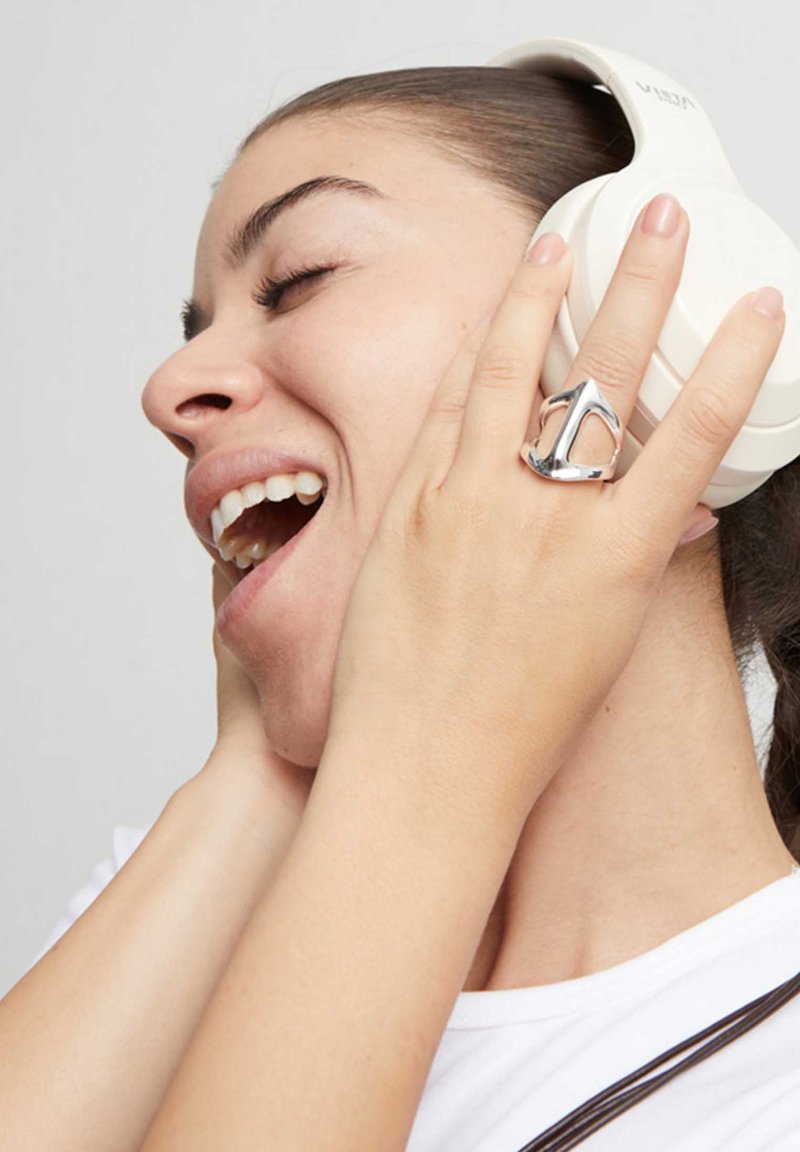 Anillo de plata con diseño puntiagudo en una mano, auriculares blancos de diadema en la cabeza de una mujer, que lleva una camiseta blanca lisa.