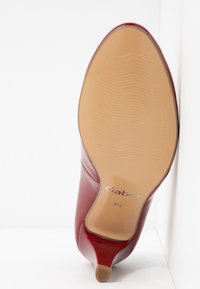 Scarpa con tacco alto in pelle lucida rossa, con punta appuntita, caratterizzata da una finitura liscia e brillante e una suola beige testurizzata con dettaglio del logo.