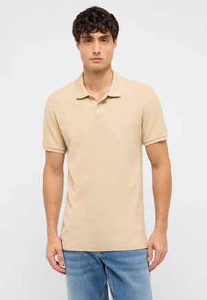 Junger Mann mit lockigem Haar, der ein beige-farbenes Poloshirt mit kurzen Ärmeln und blaue Jeans trägt und vor einem schlichten weißen Hintergrund steht.