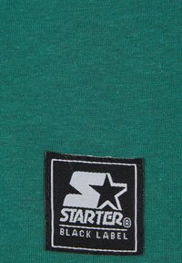 Zöld szövet, sima textúrával, fekete téglalap alakú címkével, fehér betűkkel és csillaglogóval, amelyen az áll: "STARTER BLACK LABEL."