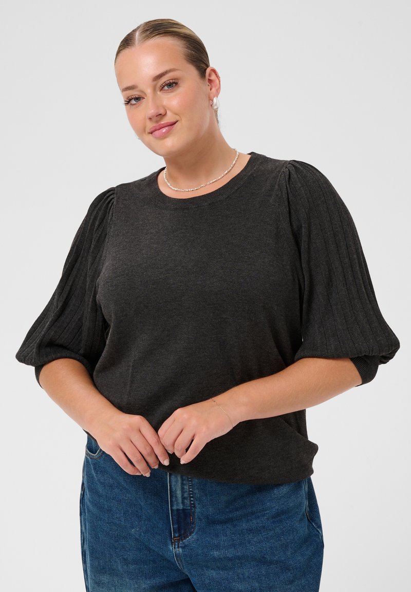 Femme aux cheveux plaqués en arrière, portant un pull gris foncé à manches côtelées bouffantes et un jean bleu sur un fond clair uni.
