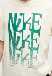 Μπεζ μπλουζάκι με μεγάλο πράσινο pixel-γραφή "NIKE" επαναλαμβανόμενο κάθετα και μικρότερο κείμενο "NIKE INC." και "OREGON, USA" στο κάτω μέρος.