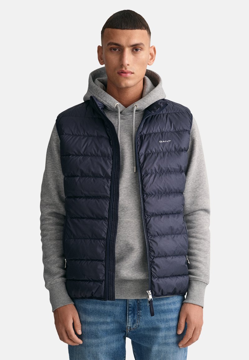 Chaleco acolchado navy con cremallera en la parte delantera, costuras horizontales y detalle de logo, llevado sobre una sudadera gris con capucha. Unos jeans de denim casuales completan el look.