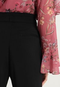 Pantalones negros de cintura alta con un frente plano y bolsillo, combinados con una blusa floral rosa transparente con mangas acampanadas y patrones vibrantes.