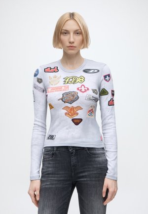 Donna con capelli biondi corti che indossa una maglietta a maniche lunghe grigio chiaro coperta da loghi colorati in stile toppa e jeans grigio scuro.