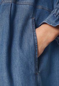 Main insérée dans la poche latérale d'un vêtement en denim bleu avec des surpiqûres orange visibles et un poignet de manche froncé.