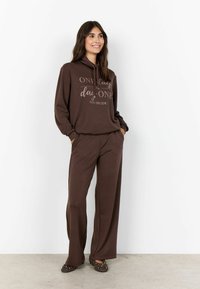 Brown hoodie with printed text, paired with matching wide-leg pants. Soft material and relaxed fit, complemented by dotted slip-on shoes.
