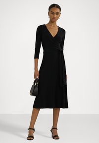 Black wrap dress with three-quarter sleeves, a fitted waist, and flared skirt. Smooth texture and a side tie detail. Paired with black heels and handbag.